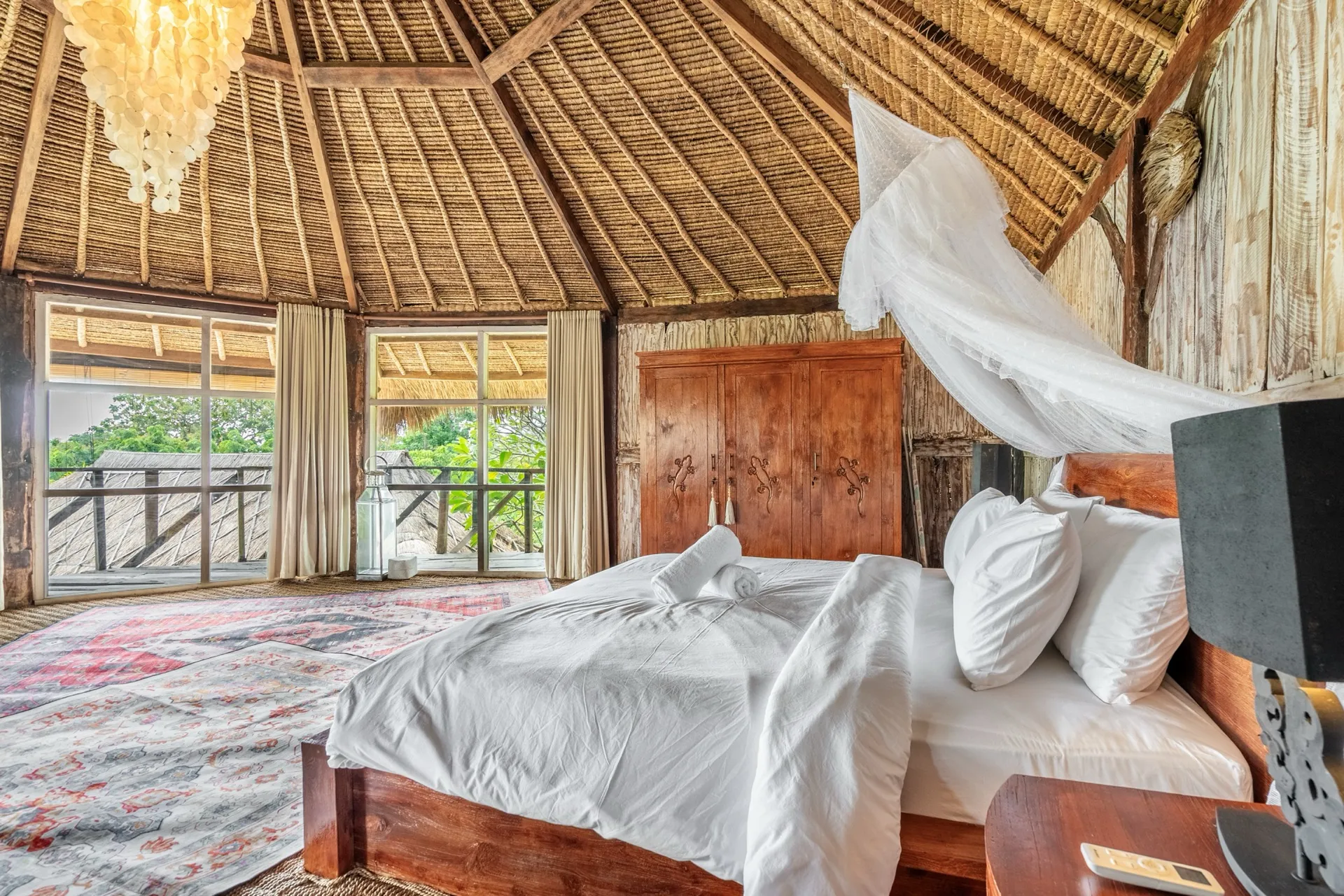 A bedroom suite at Villa Idaman beneath a high thatched ceiling, with a canopy net, carved teak wardrobe and balcony over the garden