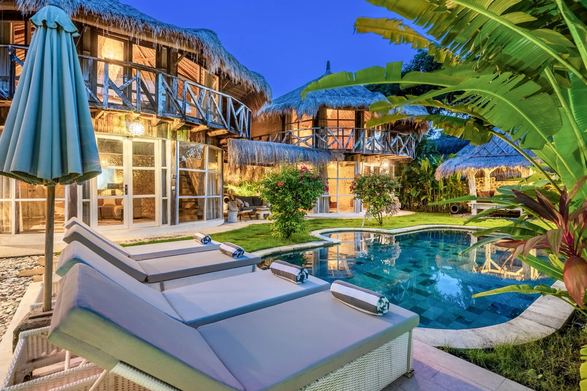 Villa Idaman's pool and sun loungers at dusk, the thatched pavilions glowing warmly behind the garden
