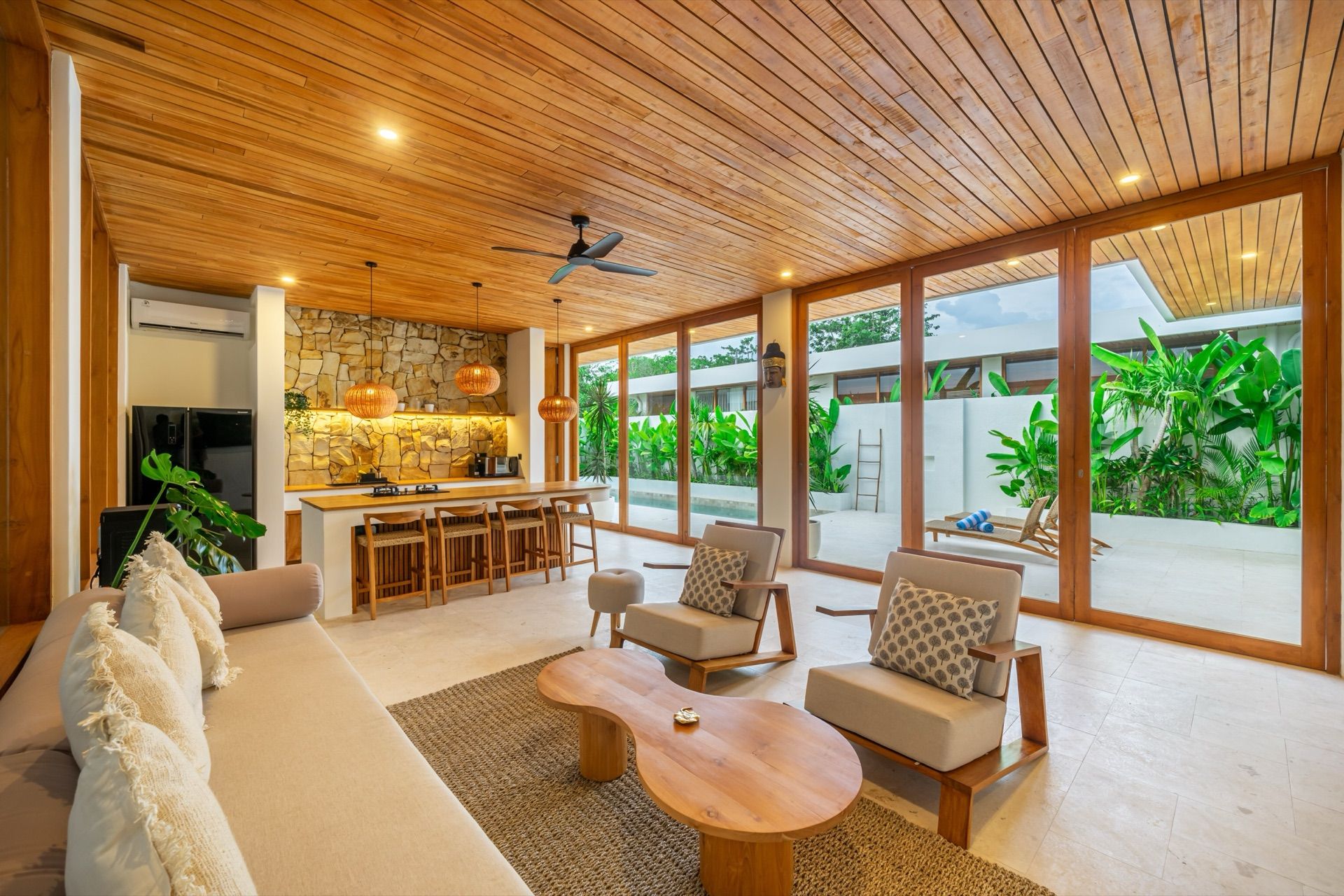 Villa Lila's open-plan living room and kitchen bar under a timber ceiling, with glass walls sliding open to the pool deck