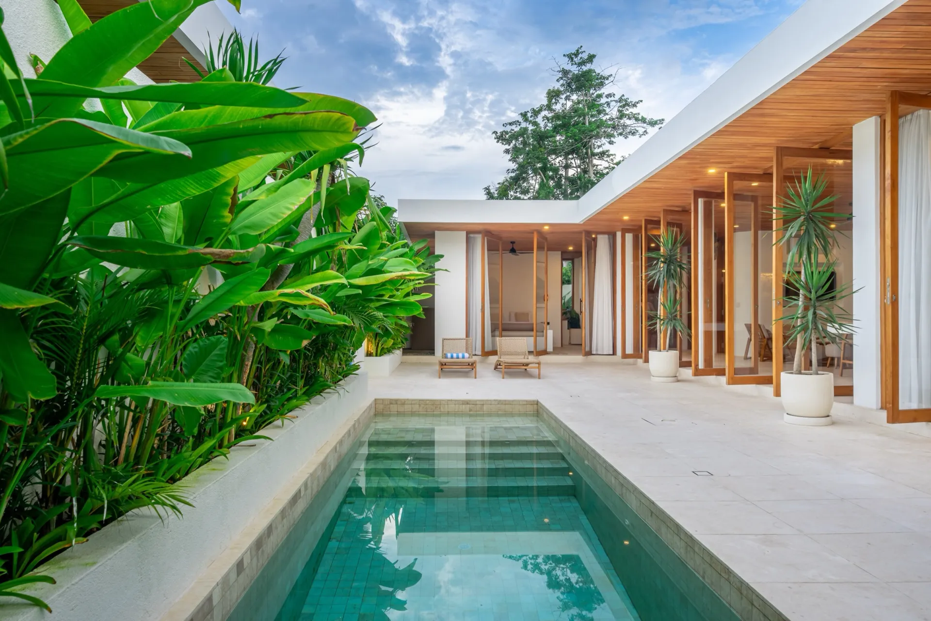 Villa Lila's private pool running the length of the terrace at dusk, bordered by dense tropical planting on one side and the villa's timber-lined pavilions on the other