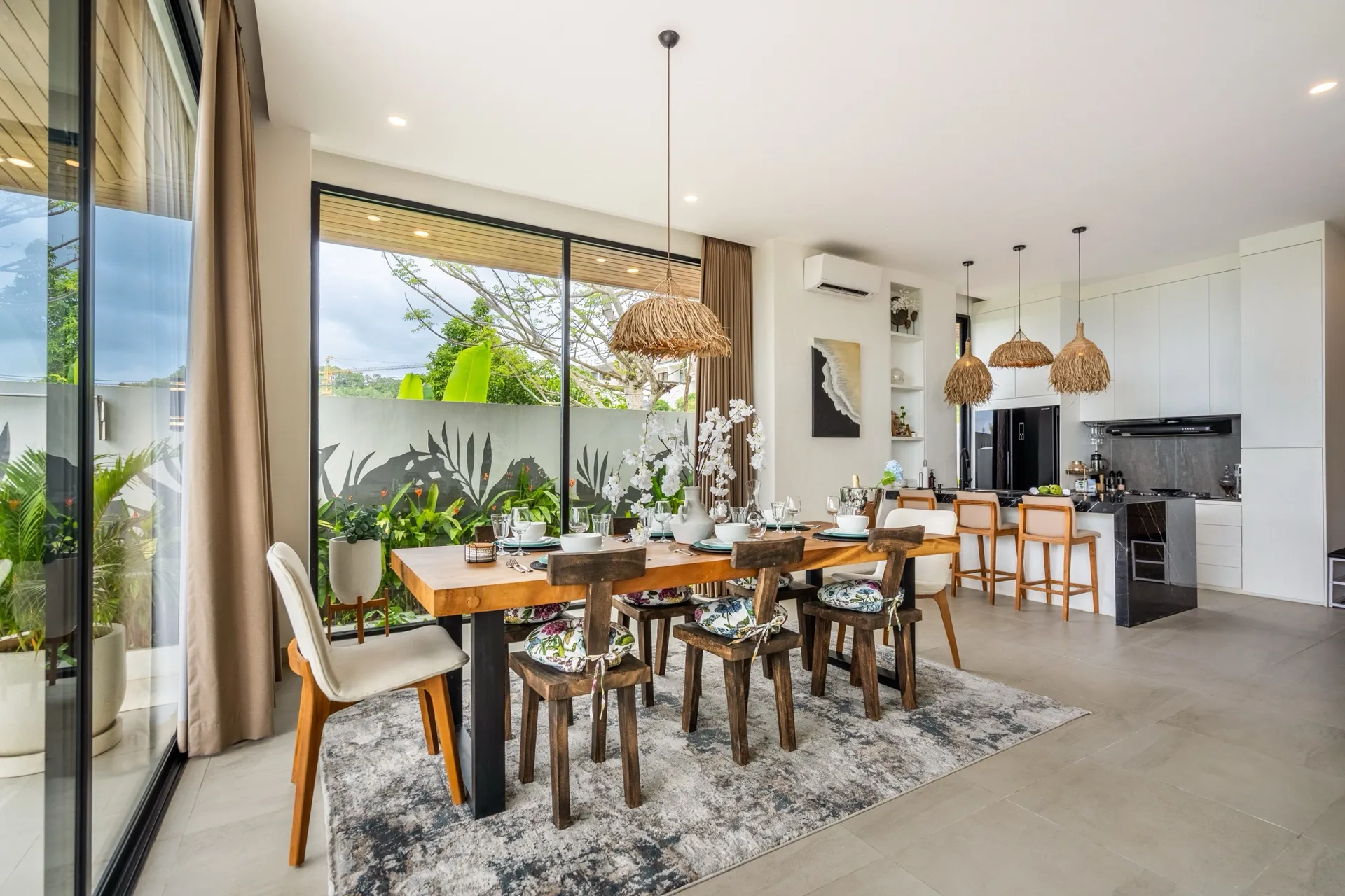 Villa Pepper's dining table in live-edge timber under raffia pendants, beside the black stone kitchen island
